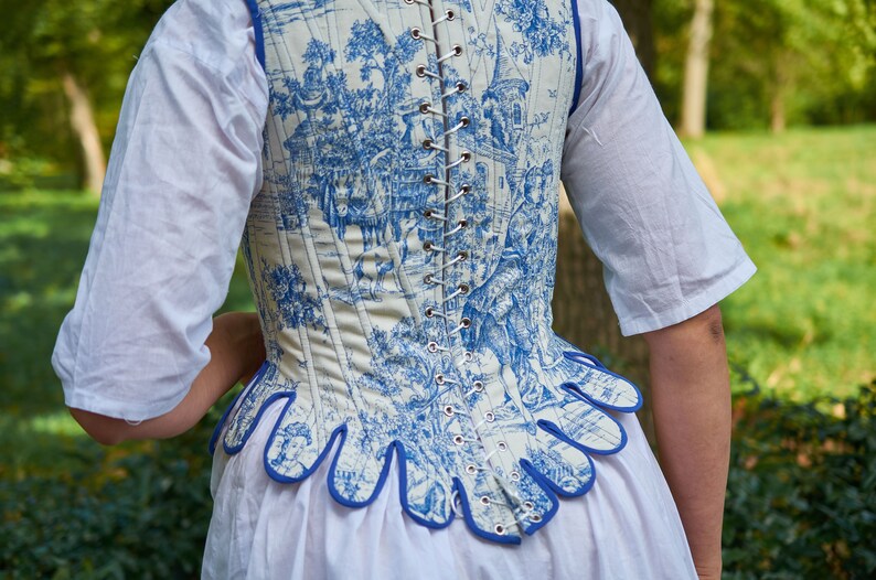 May include: A white corset with a blue and white toile pattern. The corset has a blue trim and is laced up the back.