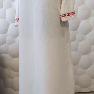 Könnte beinhalten: Weißes Leinenkleid mit roter und goldener Stickerei. Das Kleid hat ein dekoratives Muster um den Halsausschnitt, die Ärmel und den Saum. Das Kleid wird auf einer Schaufensterpuppe präsentiert.