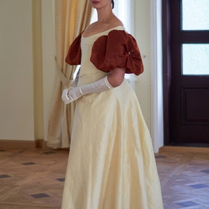 May include: A woman in a white gown with large, puffed, rust-colored sleeves stands in a room with wood floors. She is wearing white gloves and a pearl headband.