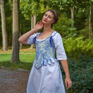 May include: A woman wearing a white dress with a blue and white patterned corset. The corset has a scalloped hem and blue ribbon ties. She is standing in a forest setting.