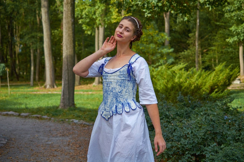 May include: A woman wearing a white dress with a blue and white patterned corset. The corset has a scalloped hem and blue ribbon ties. She is standing in a forest setting.