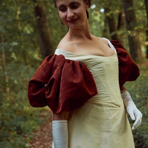 May include: A woman in a white dress with large, puffed, burgundy sleeves stands in a forest. The dress has a fitted bodice and a full skirt.
