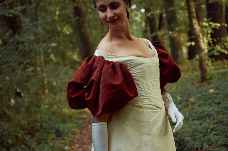 May include: A woman in a white dress with large, puffed, burgundy sleeves stands in a forest. The dress has a fitted bodice and a full skirt.