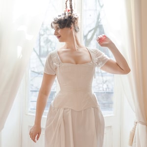 May include: A woman wearing a white corset and a white skirt with a large, white bow in the back. She has her hair styled in a high bun with a red ribbon.