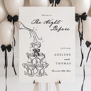 May include: A white sign with black text and a black and white illustration of champagne glasses. The text reads "Welcome to The Night Before celebrating ADELENE and THOMAS December 20th, 2020". The sign is propped up on an easel in front of a white wall with a balloon arch made of white balloons with black ribbon bows.