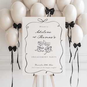 Könnte beinhalten: Ein Schild für eine Verlobungsfeier auf einer Staffelei, umgeben von weißen Luftballons mit schwarzen Schleifen. Auf dem Schild steht "Welcome to Adelene + Thomas's ENGAGEMENT PARTY" mit Datum und Toast-Nachricht. Der Hintergrund ist eine weiße Wand.