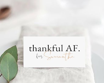 May include: A white place card with the text "thankful AF. for Samantha" in black and gold lettering. The place card is on a beige linen napkin on a white plate with a sprig of greenery.