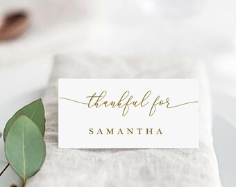May include: White place card with gold calligraphy that reads "thankful for" and the name "Samantha". The place card is on a folded linen napkin on a white plate.