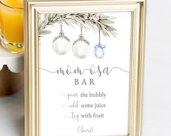 May include: A gold-framed sign with the text "Mom-osa Bar" and instructions. Three glasses of orange juice sit on a white cake stand, with a floral arrangement in the background. The sign has watercolor ornaments.