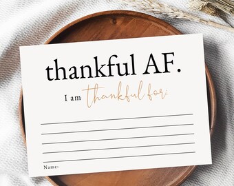 Puede incluir: Una tarjeta blanca imprimible con el texto "thankful AF. I am thankful for:" y líneas en blanco para escribir. La tarjeta está sobre un plato de madera con un fondo blanco.