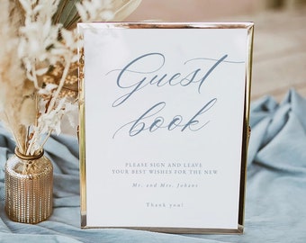 May include: A gold-framed sign with a white background and blue text that reads "Guest book". The sign also includes the text "Please sign and leave your best wishes for the new Mr. and Mrs. Johans. Thank you!" and the logo "Averly Paperie".