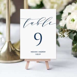 May include: White table number card with a navy blue cursive "table" and the number "9" in navy blue. The card is on a small wooden easel. The text "MEGHAN + ANDREW 9.20.19" is printed in navy blue at the bottom of the card.