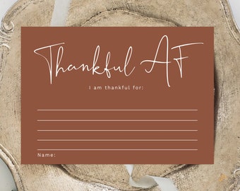Puede incluir: Una tarjeta de color terracota con las palabras "Thankful AF" en escritura blanca. La tarjeta incluye indicaciones para escribir por qué estás agradecido y un espacio para un nombre. La tarjeta está sobre un plato decorativo.