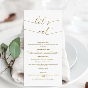 May include: A formal menu card with the words "let's eat" in elegant gold script. The menu lists a multi-course meal, including soup, salad, turkey, and dessert. The card is set on a white plate with copper-colored silverware.