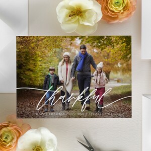 Puede incluir: Una familia de cuatro personas camina por un sendero en un bosque con follaje oto&ntilde;al. Los padres van de la mano con sus dos hijos. El texto "thankful" est&aacute; escrito en cursiva blanca en la parte inferior de la imagen. El texto "Happy Thanksgiving 2021 | The Andersons" est&aacute; escrito en blanco debajo de la cursiva.