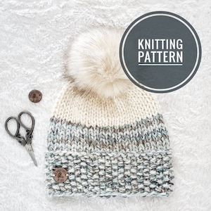 Peut inclure: Un bonnet tricoté avec un pompon blanc, une bande rayée blanche et grise et un motif moucheté bleu et gris. Le bonnet est doté d'une étiquette en cuir marron avec le texte "Embroidered by You".