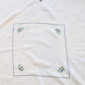 May include: Cream tablecloth with embroidered details. The top edge has a row of colourful cross-stitched designs, including a pagoda and foliage. The edges are finished with a dark blue zigzag stitch, and the centre has a square with four embroidered motifs.