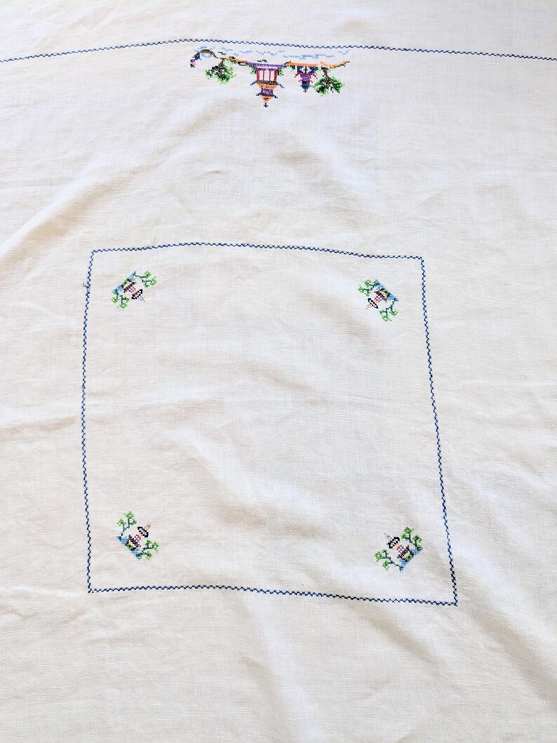 May include: Cream tablecloth with embroidered details. The top edge has a row of colourful cross-stitched designs, including a pagoda and foliage. The edges are finished with a dark blue zigzag stitch, and the centre has a square with four embroidered motifs.