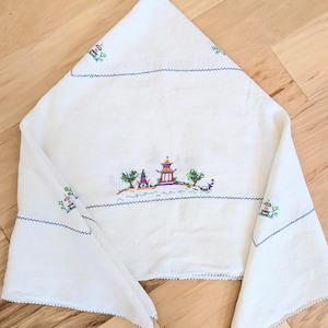 May include: White linen cloth featuring a cross-stitched Asian landscape. The embroidery depicts a pagoda, trees, and a boat, with blue stitching outlining the edges and details. The cloth is folded, showcasing the design.