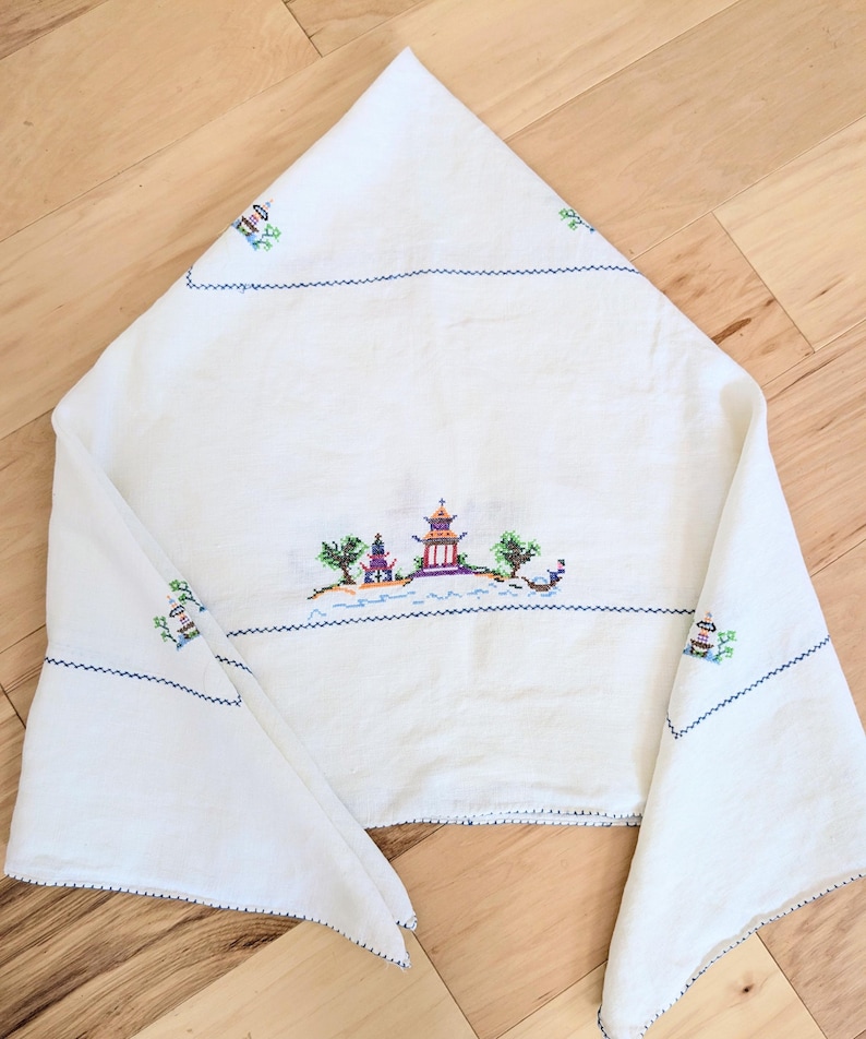 May include: White linen cloth featuring a cross-stitched Asian landscape. The embroidery depicts a pagoda, trees, and a boat, with blue stitching outlining the edges and details. The cloth is folded, showcasing the design.