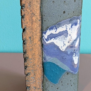 May include: A triangular ceramic vase with a textured, dark brown vertical edge and a smooth, multi-colored face featuring shades of gray, blue, and white. The surface displays a mottled glaze with a distinct white drip pattern and a circular opening.