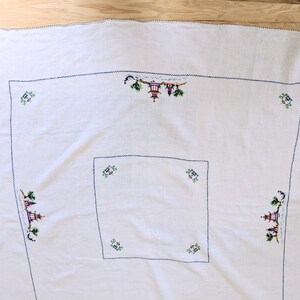 May include: White tablecloth with embroidered Asian-inspired scenes in green, purple, and yellow. The embroidery features pagodas and trees, outlined with a dark blue stitch. The tablecloth has a square design with a smaller square in the centre.