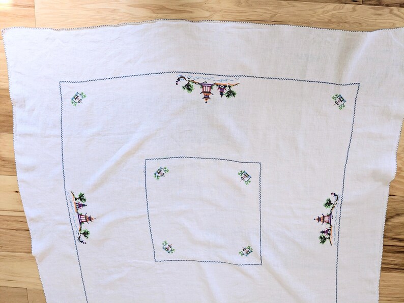 May include: White tablecloth with embroidered Asian-inspired scenes in green, purple, and yellow. The embroidery features pagodas and trees, outlined with a dark blue stitch. The tablecloth has a square design with a smaller square in the centre.