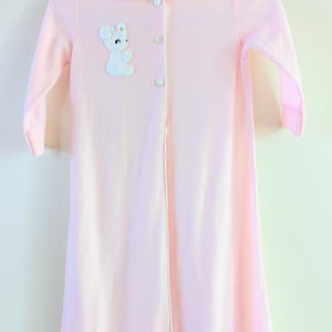 May include: A light pink baby gown with long sleeves, a Peter Pan collar, and a white bear applique. The gown has three buttons down the front and a white trim along the collar and front opening. The gown is hanging on a white hanger.