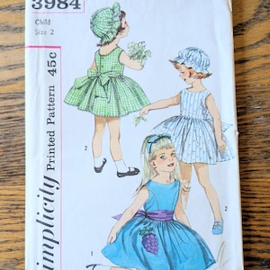 May include: A Simplicity sewing pattern envelope 3984 for a child size 2 dress. The illustration shows three children wearing sleeveless dresses and bonnets. One dress is green gingham, one is blue striped, and one is solid blue with a purple sash and grapes.