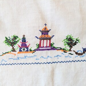 May include: A cross-stitch embroidery on white fabric shows a colourful Asian-inspired landscape. The scene includes pagodas, trees, and a boat, all rendered in shades of purple, orange, green, and blue. The design is framed by a blue border.