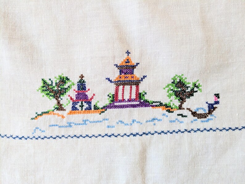 May include: A cross-stitch embroidery on white fabric shows a colourful Asian-inspired landscape. The scene includes pagodas, trees, and a boat, all rendered in shades of purple, orange, green, and blue. The design is framed by a blue border.