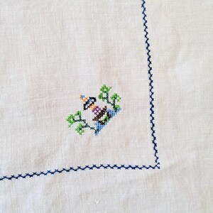 May include: White linen tablecloth with a detailed cross-stitch design in the corner. The cross-stitch features a lighthouse with green trees and blue water. A blue stitched border frames the edge.