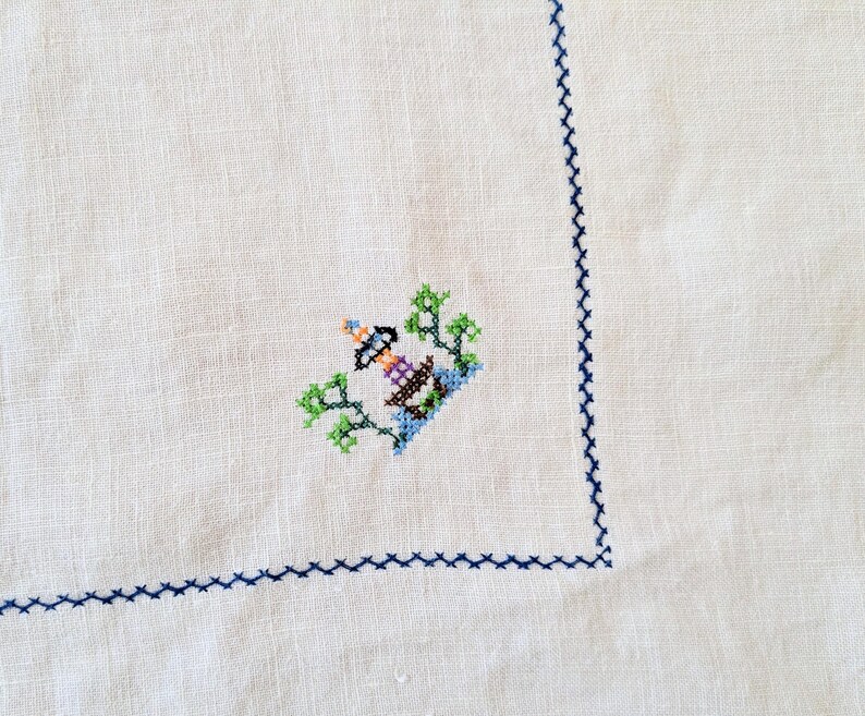 May include: White linen tablecloth with a detailed cross-stitch design in the corner. The cross-stitch features a lighthouse with green trees and blue water. A blue stitched border frames the edge.