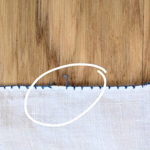 May include: Close-up of white fabric with a blue stitched border. The fabric is on a wooden surface. The blue stitching creates a decorative edge along the bottom of the fabric.