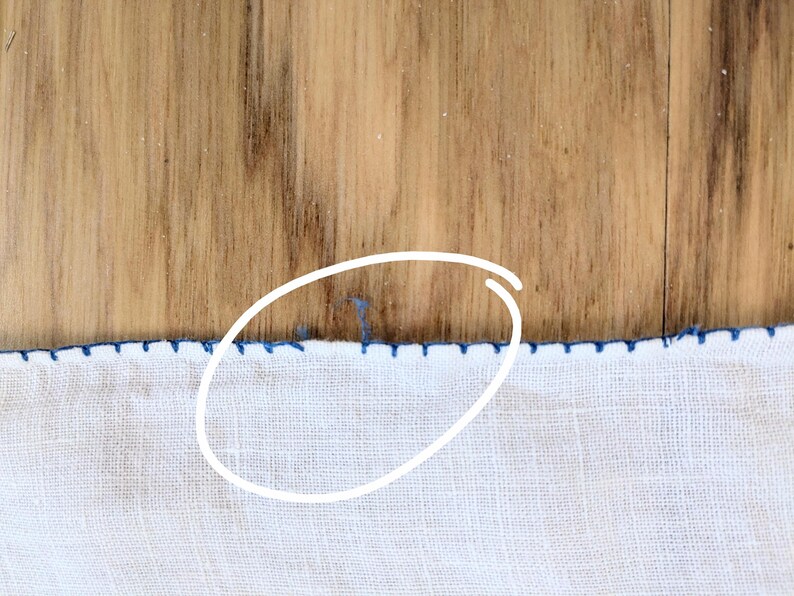 May include: Close-up of white fabric with a blue stitched border. The fabric is on a wooden surface. The blue stitching creates a decorative edge along the bottom of the fabric.