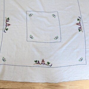 May include: White tablecloth with embroidered Asian landscape scenes in blue, green, purple, and red. The tablecloth has a square centre and is bordered with a blue stitched edge.
