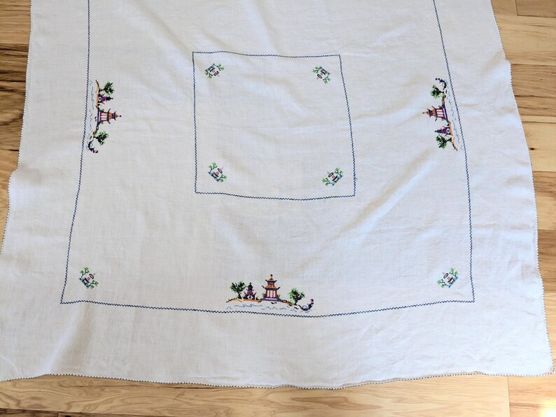 May include: White tablecloth with embroidered Asian landscape scenes in blue, green, purple, and red. The tablecloth has a square centre and is bordered with a blue stitched edge.