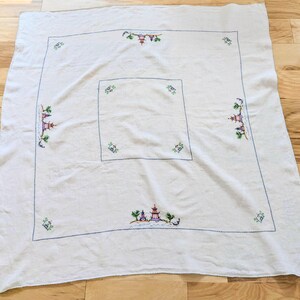 May include: A white tablecloth featuring a blue stitched border and embroidered Asian-inspired pagoda and floral designs. The tablecloth is square with a smaller square design in the centre.