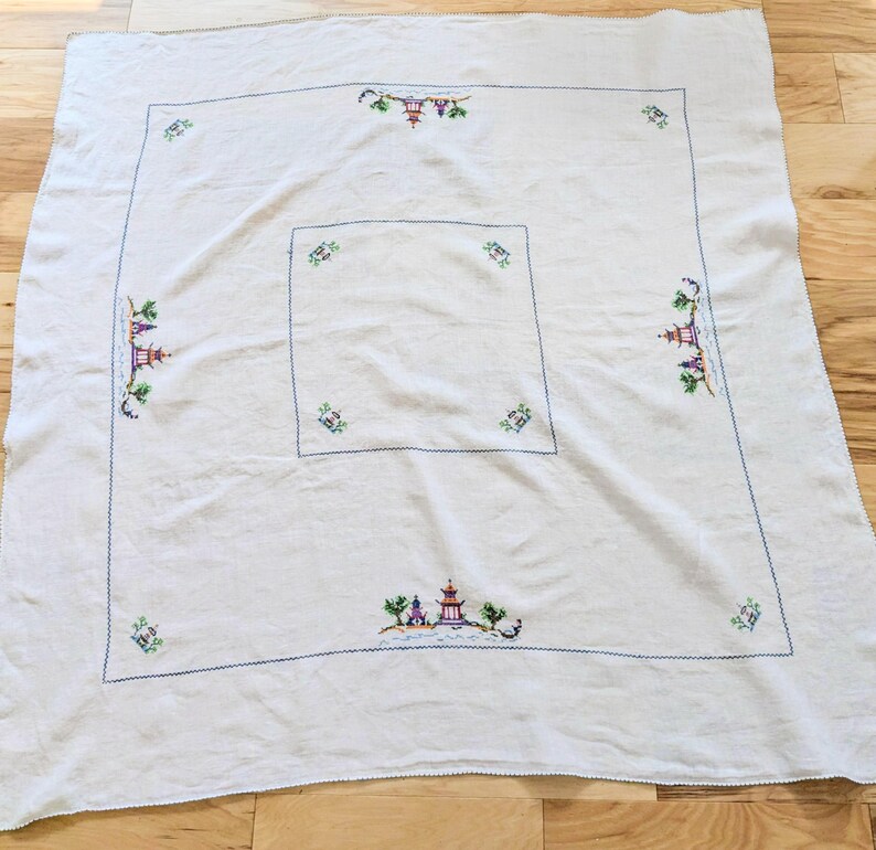 May include: A white tablecloth featuring a blue stitched border and embroidered Asian-inspired pagoda and floral designs. The tablecloth is square with a smaller square design in the centre.