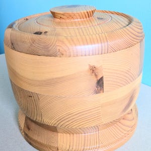 May include: A light-colored wooden container with a lid. The container is cylindrical with a rounded base and a lid with a small knob. The wood grain is visible throughout the piece, and the container appears to be handcrafted.