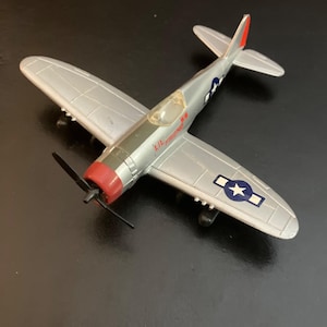 May include: A silver toy airplane with a red nose, black propeller, and landing gear. The fuselage features the text LIL FRIEND in red lettering, and the wings display blue and white star insignias with rectangular bars on each side.