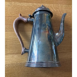 Puede incluir: Una cafetera plateada con un asa de madera y un pico curvo. La cafetera tiene una tapa con un pequeño pomo en la parte superior. El asa es de color marrón claro y se curva alejándose del cuerpo de la cafetera. Aproximadamente 25 cm de altura.