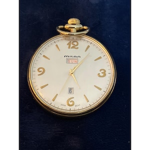 May include: A gold-toned Milan pocket watch with a cream-colored face. The watch features gold numerals, hands, and a date window at the 6 o'clock position. The word "SUN" is displayed above the hands. The watch is marked "Japan" and "MCVT".