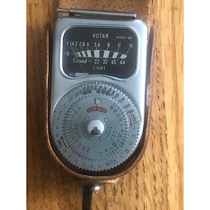 May include: A vintage VOTAR Hyper-III light meter in a brown leather case. The meter has a silver face with black text and a dial for light value and time settings. The meter reads "VOTAR" and "HYPER-III".