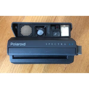 May include: A black Polaroid Spectra AF instant camera. The camera has a built-in flash, a lens, and the word "Polaroid" printed on the front. The camera also has the words "Spectra AF" printed on the front.