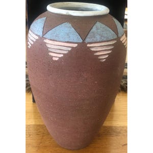 May include: A large, brown ceramic vase with a wide base and a narrow neck. The vase is decorated with geometric shapes in light blue and pink. The rim is white.