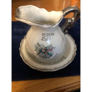 Pode incluir: Conjunto de jarro de creme e açucareiro de porcelana branca com detalhes prateados. O jarro de creme apresenta um design de 25º aniversário com sinos e flores. O açucareiro tem uma borda festonada.