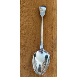 May include: A silver spoon with a long, slender handle and a rounded bowl. The handle has a rectangular top. The spoon is highly polished, reflecting the surrounding environment. The spoon is likely for serving or eating.