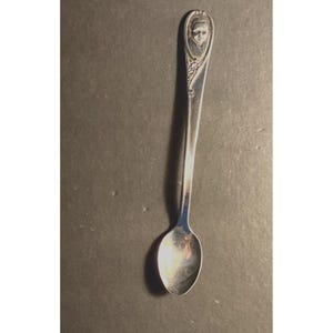 Könnte beinhalten: Ein silberner Löffel mit einem detaillierten Porträt am oberen Ende des Griffs. Der Löffel hat eine kleine, ovale Schüssel und einen langen, schlanken Griff. Das Porträt zeigt eine Person, und der Löffel hat eine antike oder Vintage-Ästhetik.