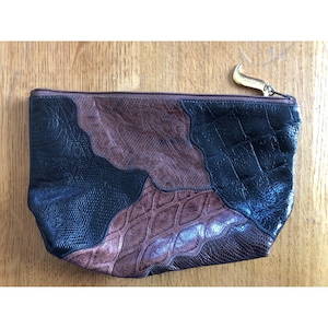 Könnte beinhalten: Eine Patchwork-Clutch mit einer Mischung aus Texturen und Farben. Das Design umfasst schwarze, braune und beige Lederstücke mit einem goldfarbenen Reißverschluss. Die Tasche hat eine rechteckige Form und einen Reißverschluss.